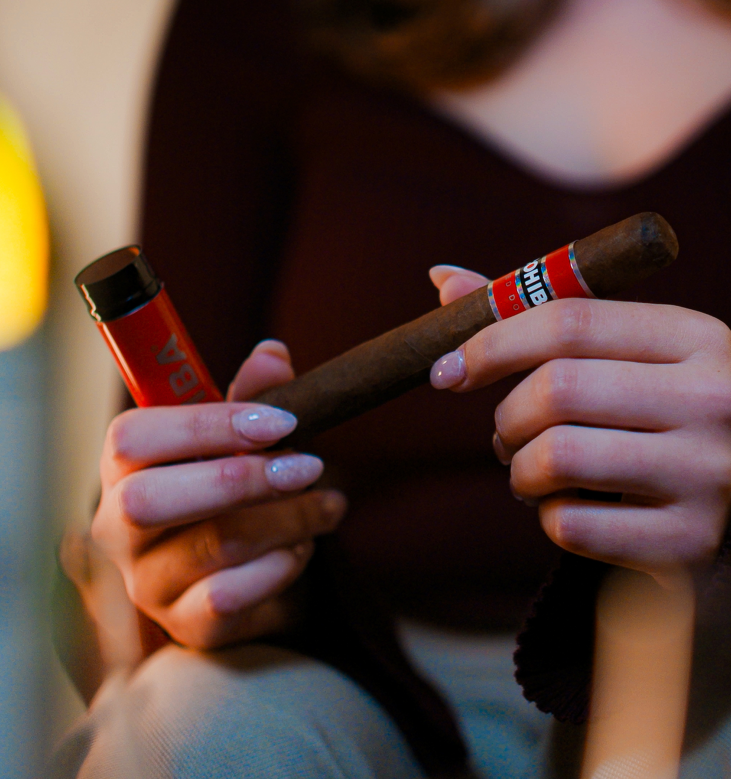 A person is holding a cigar and its case. There's blurred lighting and a bottle in the background, creating a relaxed atmosphere.