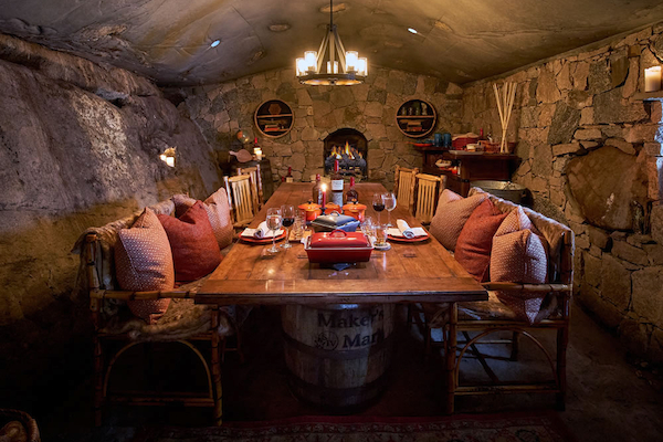 A cozy dining room with a long wooden table, stone walls, candles, and rustic decor, illuminated by a warm hanging light fixture.