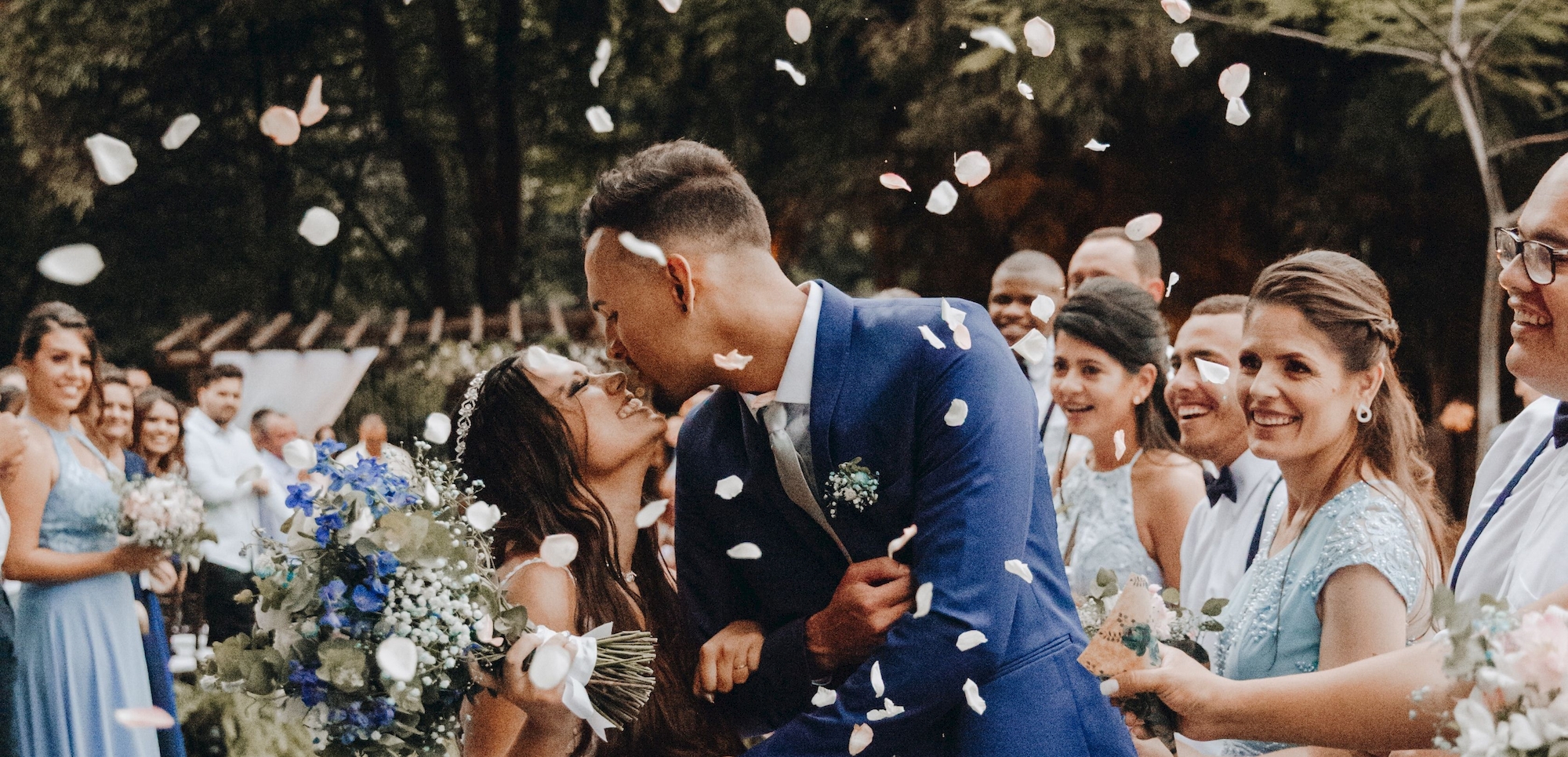 A bride and groom kiss surrounded by joyful guests and flower petals in an outdoor wedding scene, with bridesmaids in matching blue dresses smiling.