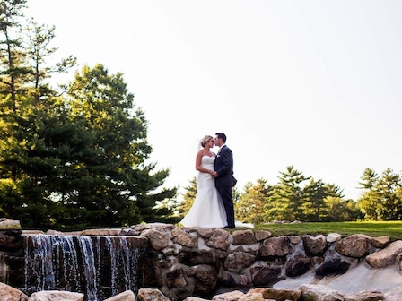 A bride and groom stand on a rock wall near a cascading waterfall, surrounded by lush greenery in an outdoor setting, embracing each other.