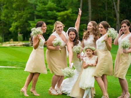 A bride, bridesmaids, and a flower girl in light dresses joyfully celebrate, holding bouquets, in a lush green outdoor setting, surrounded by trees.