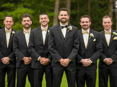 A group of six men in suits and bow ties, likely a wedding party, standing outdoors on a grassy area. Trees are visible in the background.