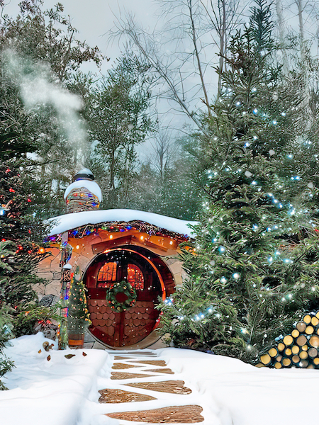 A cozy hobbit-like house in a snowy forest, surrounded by decorated trees. Smoke rises from the chimney, and a path leads to the arched door.