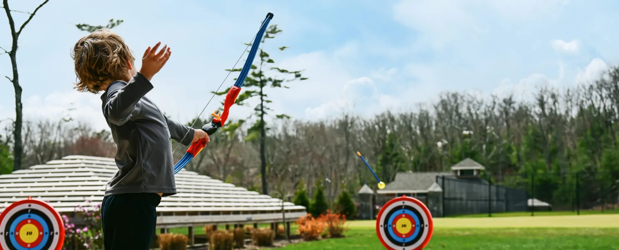 A child is using a toy bow and arrow at an outdoor archery range with colorful targets in a park on a clear day ending the sentence.