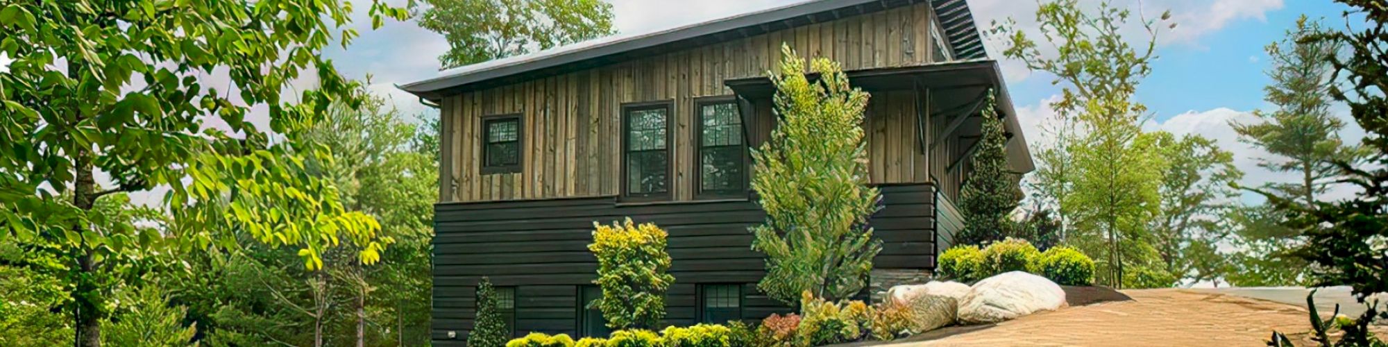 A modern wooden house with black accents sits in a lush green area, surrounded by trees and landscaped with plants and rocks, against a blue sky.