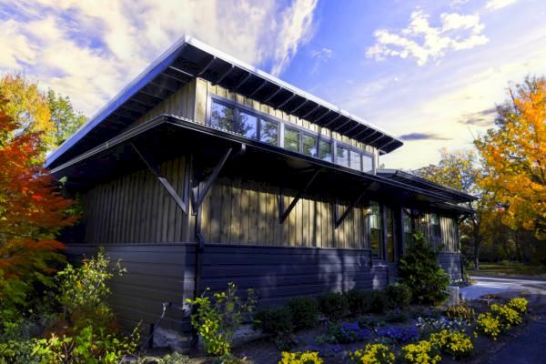 A modern building with a flat roof and large windows is surrounded by lush greenery and colorful flowers, under a vibrant sky with scattered clouds.