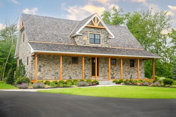 A stone house with a pitched roof, wooden beams, and a manicured lawn, surrounded by greenery and trees under a partly cloudy sky.