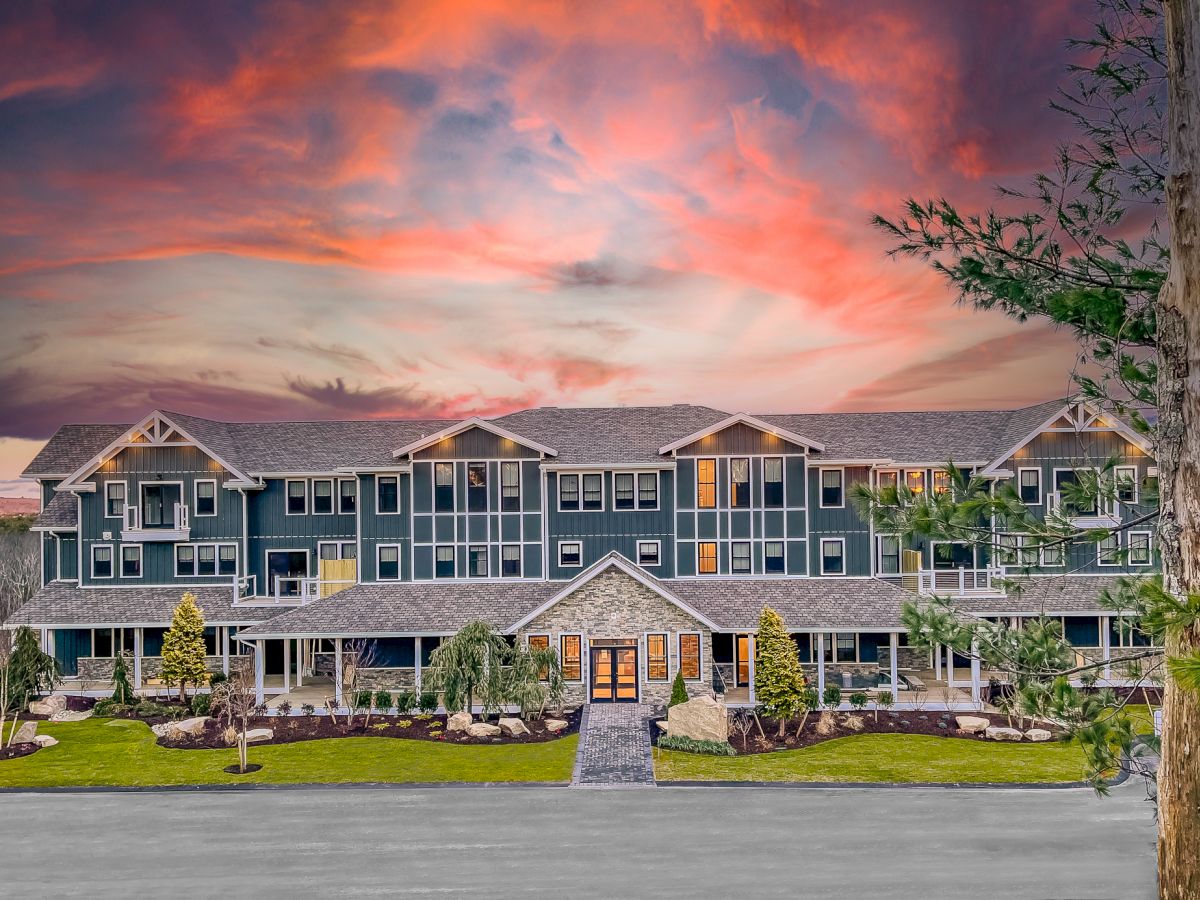 A large, modern building with multiple stories and windows, surrounded by greenery, with a dramatic sunset sky in the background.