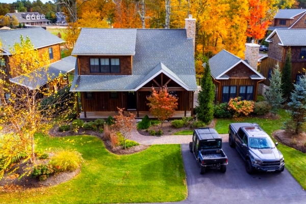 A quaint, two-story house with a well-manicured lawn, surrounded by vibrant autumn trees, and two vehicles parked in the driveway.