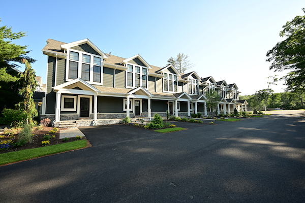 The image shows a row of modern townhouses with green lawns and a spacious driveway under a clear blue sky.