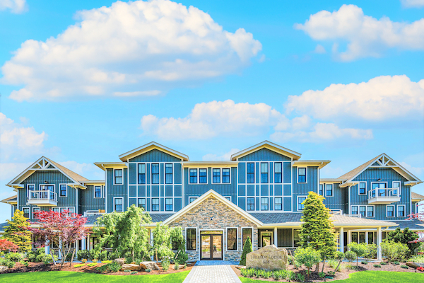 A large, modern building with blue siding, multiple windows, and a landscaped garden with a pathway leading to the entrance, under a blue sky with clouds.