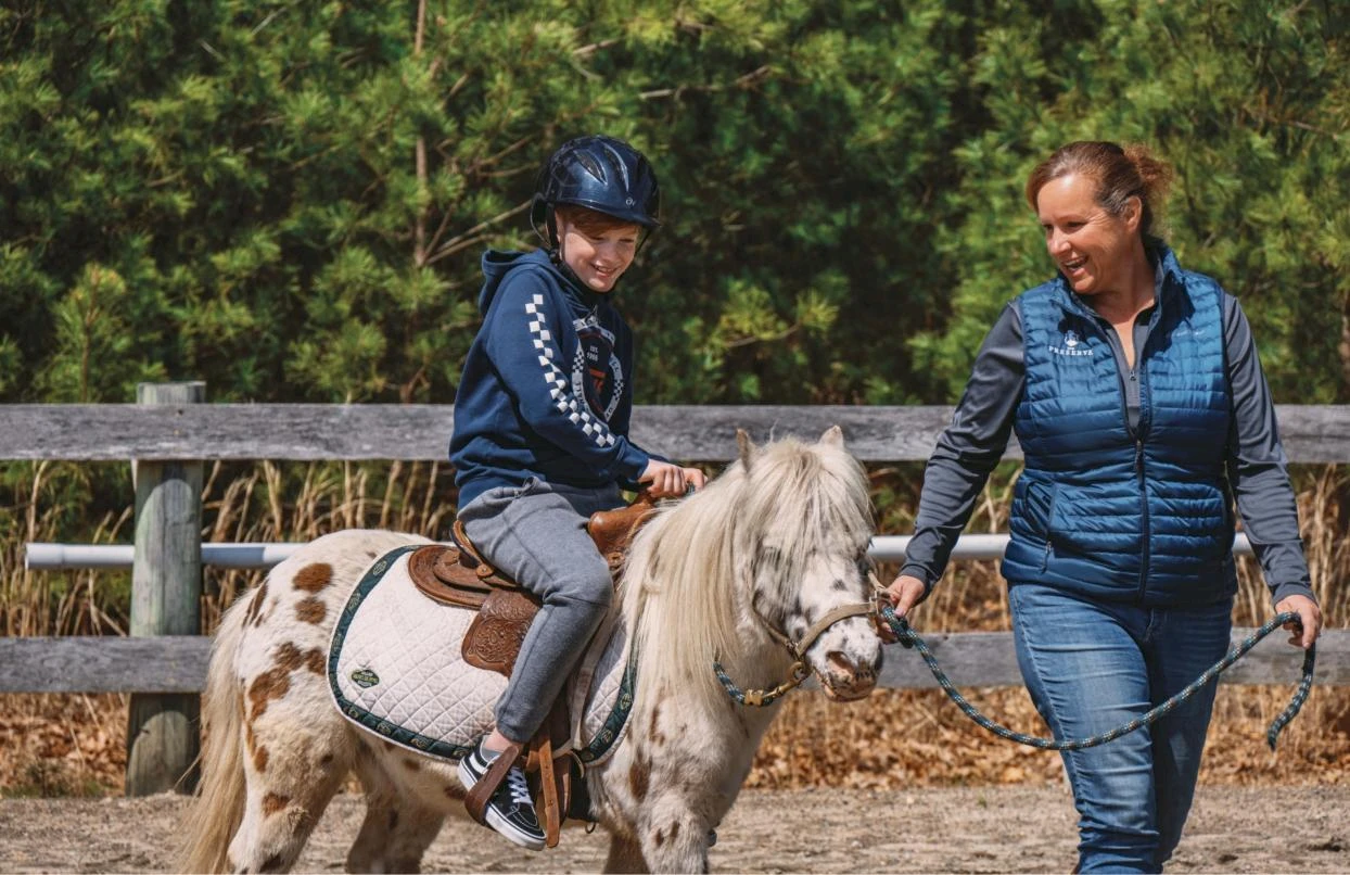 A child wearing a helmet rides a small horse while an adult holding a lead rope walks beside them in an outdoor setting.