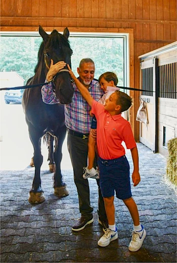 An adult and two children pet a horse inside a stable, with the adult holding one child and the other child reaching out to touch the horse.