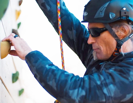 A person wearing a helmet and sunglasses climbs a wall with handholds, using a rope for safety. They appear focused on the activity.