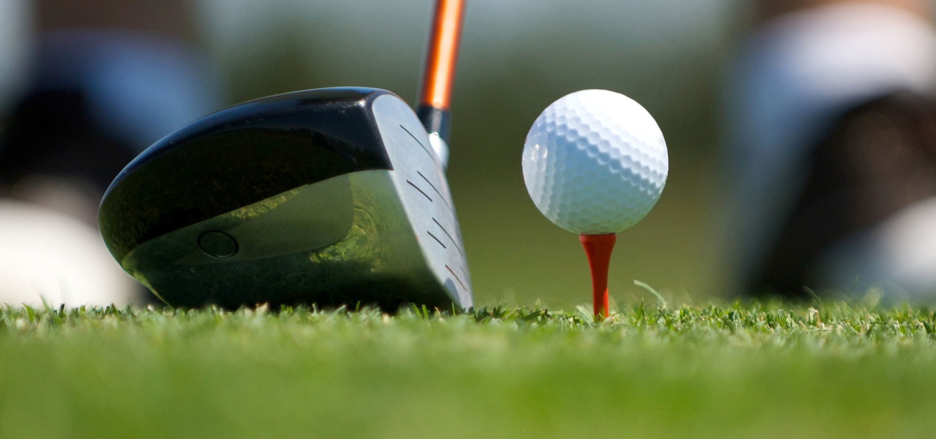 A close-up image of a golf club about to strike a golf ball on a red tee, with grass and blurred background.