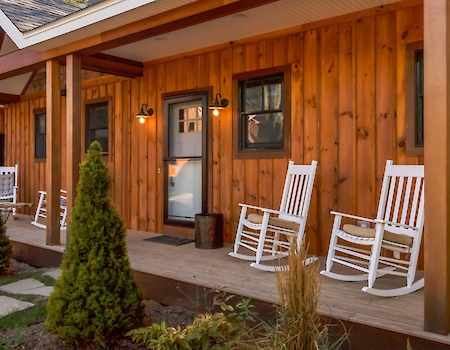 A wooden house with a porch, white rocking chairs, small trees, and a jug on the porch corner.