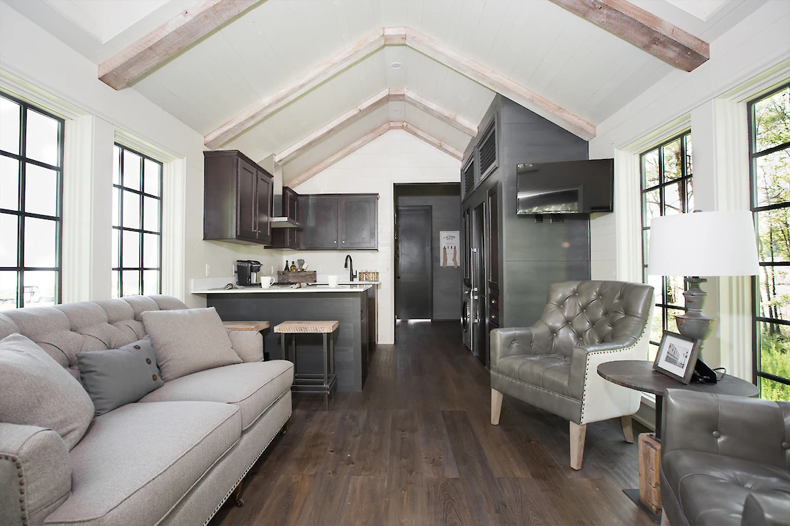 This image features the interior of a modern tiny house, showcasing a kitchen, living area with a sofa and armchair, wooden floors, and large windows.