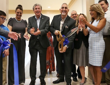 A group of people stand together at a ribbon-cutting ceremony, with one person holding large scissors and others clapping.