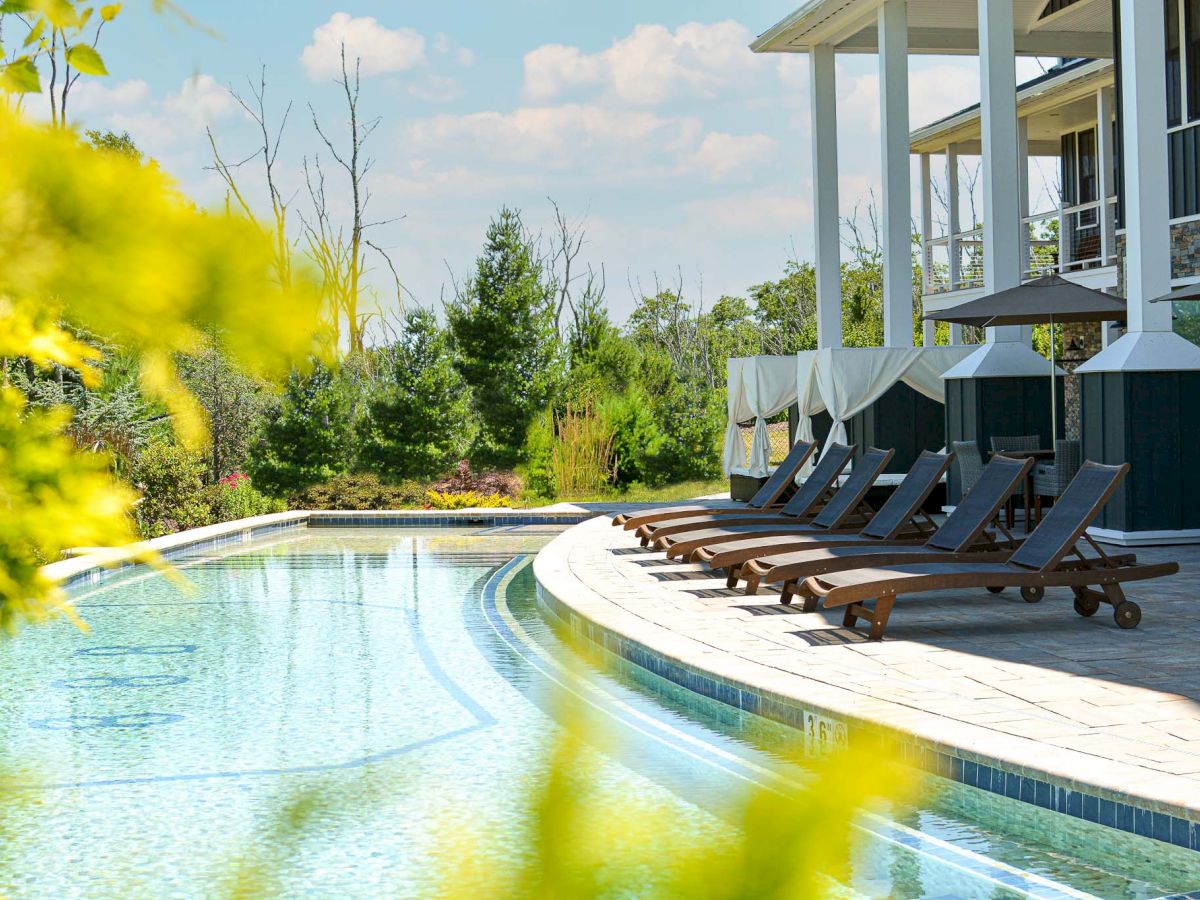 An outdoor swimming pool with several lounge chairs lined up on the deck. The area is surrounded by trees and greenery, and the sky is clear.