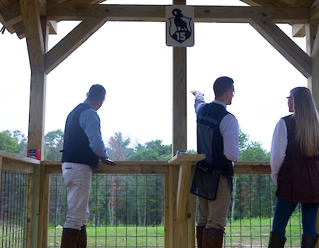 Three people in an outdoor wooden structure, seemingly at a shooting range, with one pointing towards something in the distance, ending the sentence.