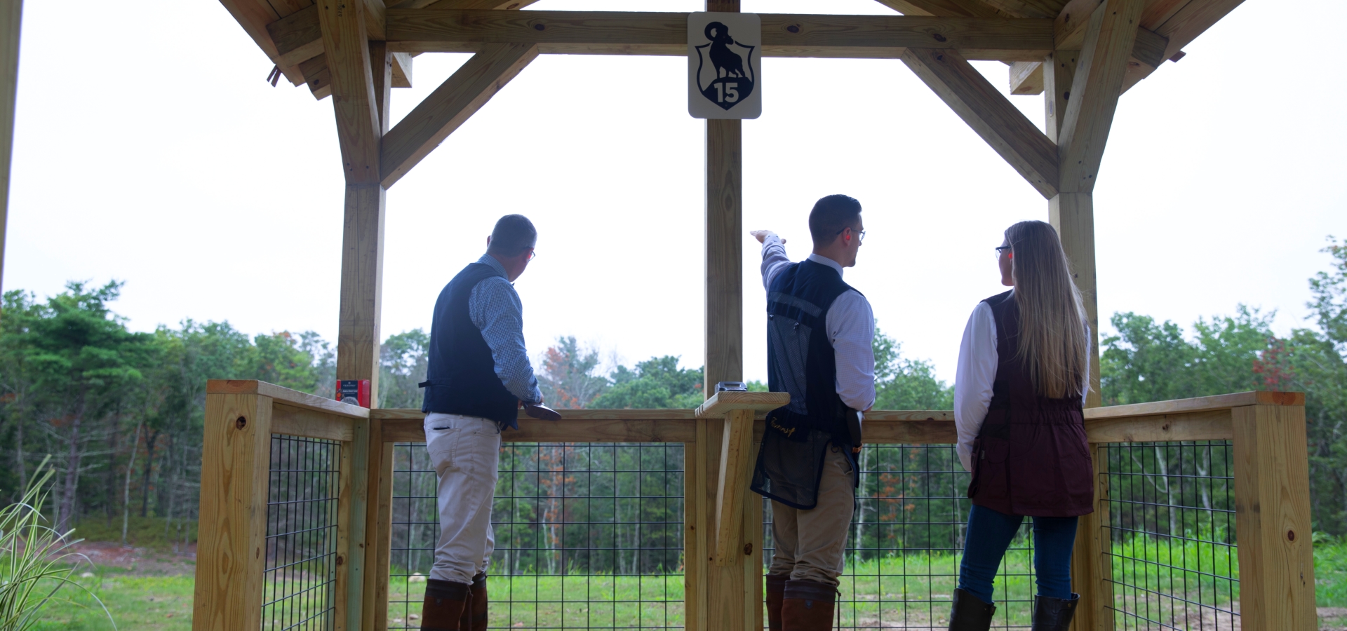 Three people in an outdoor wooden structure, seemingly at a shooting range, with one pointing towards something in the distance, ending the sentence.