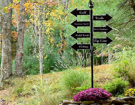 A signpost in a forested area has multiple direction signs for Golf, Trails, Mountain Sports, Fish Trails, Black & Blue Lake, Zip Line, and Fishing.