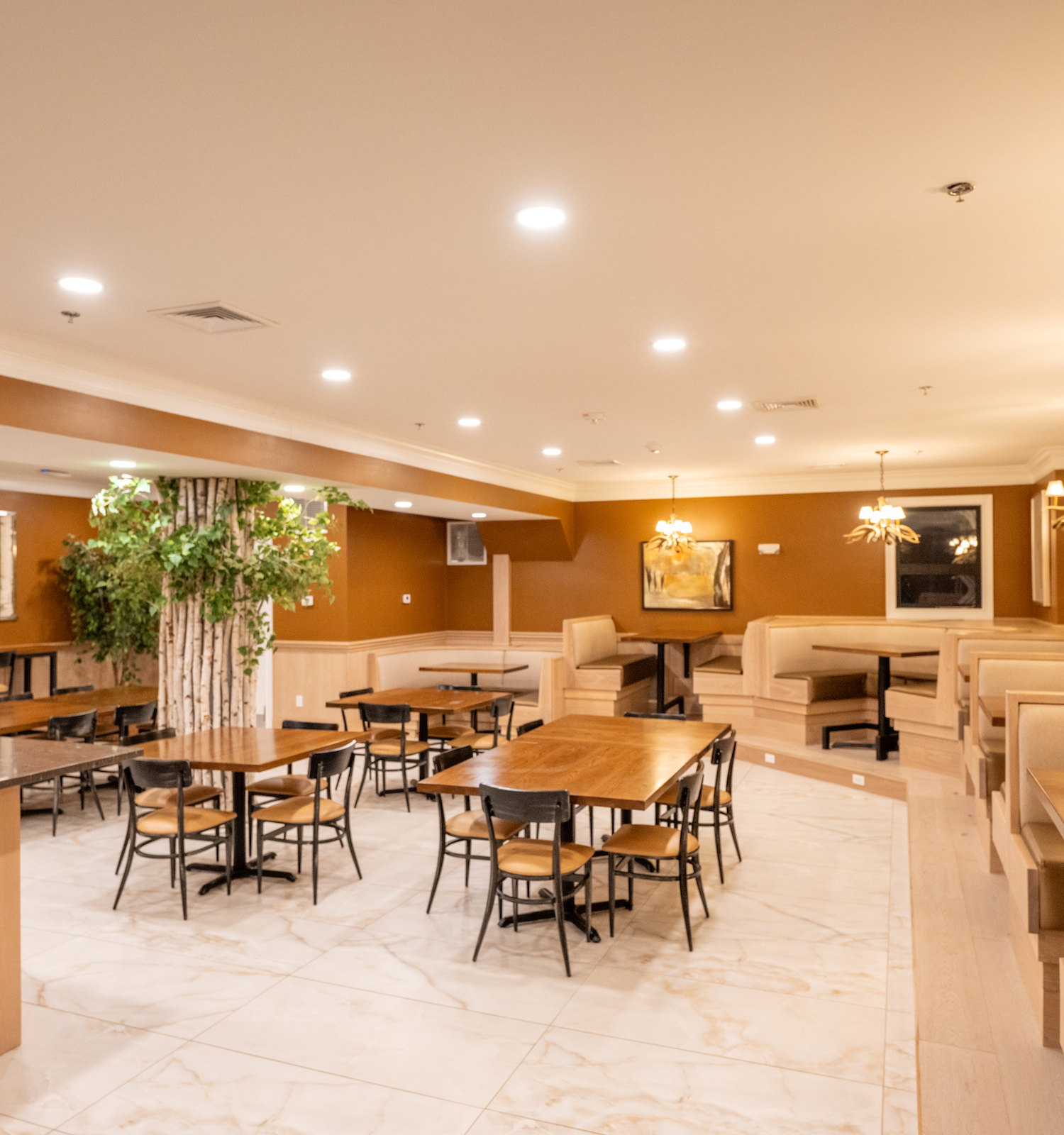 A warmly lit restaurant interior with tables, chairs, and booths arranged in a spacious dining area, adorned with decorative plants.