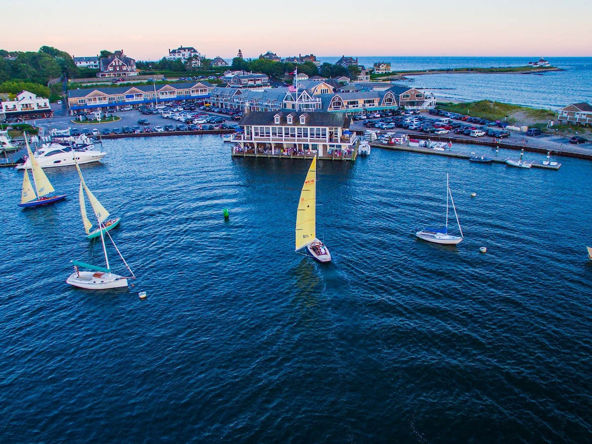 Aerial view of a marina with boats on the water, surrounded by buildings and greenery, with a coastline and open sea in the background.