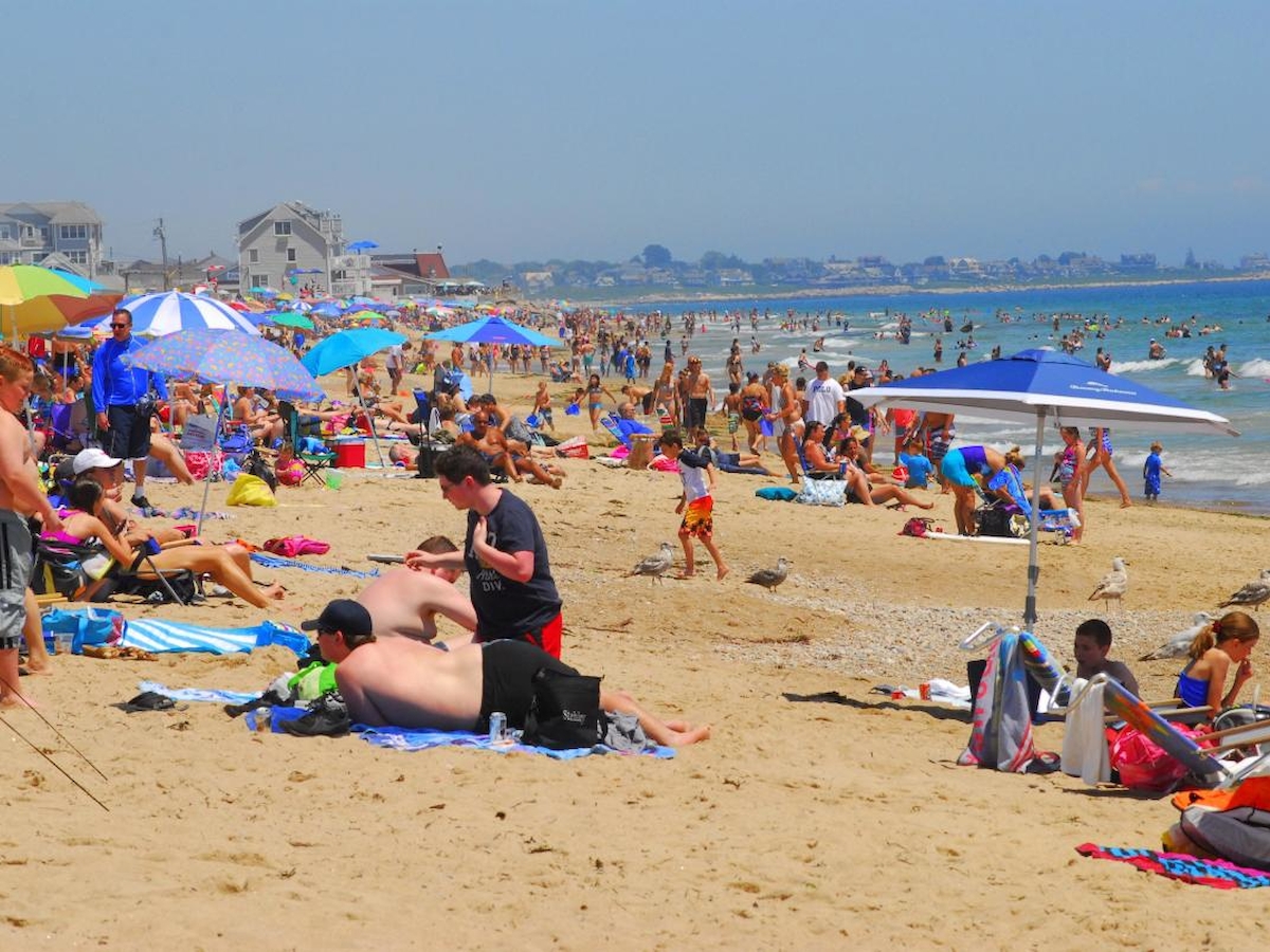 The image shows a crowded beach with people sunbathing, swimming, and relaxing under umbrellas on a sunny day.