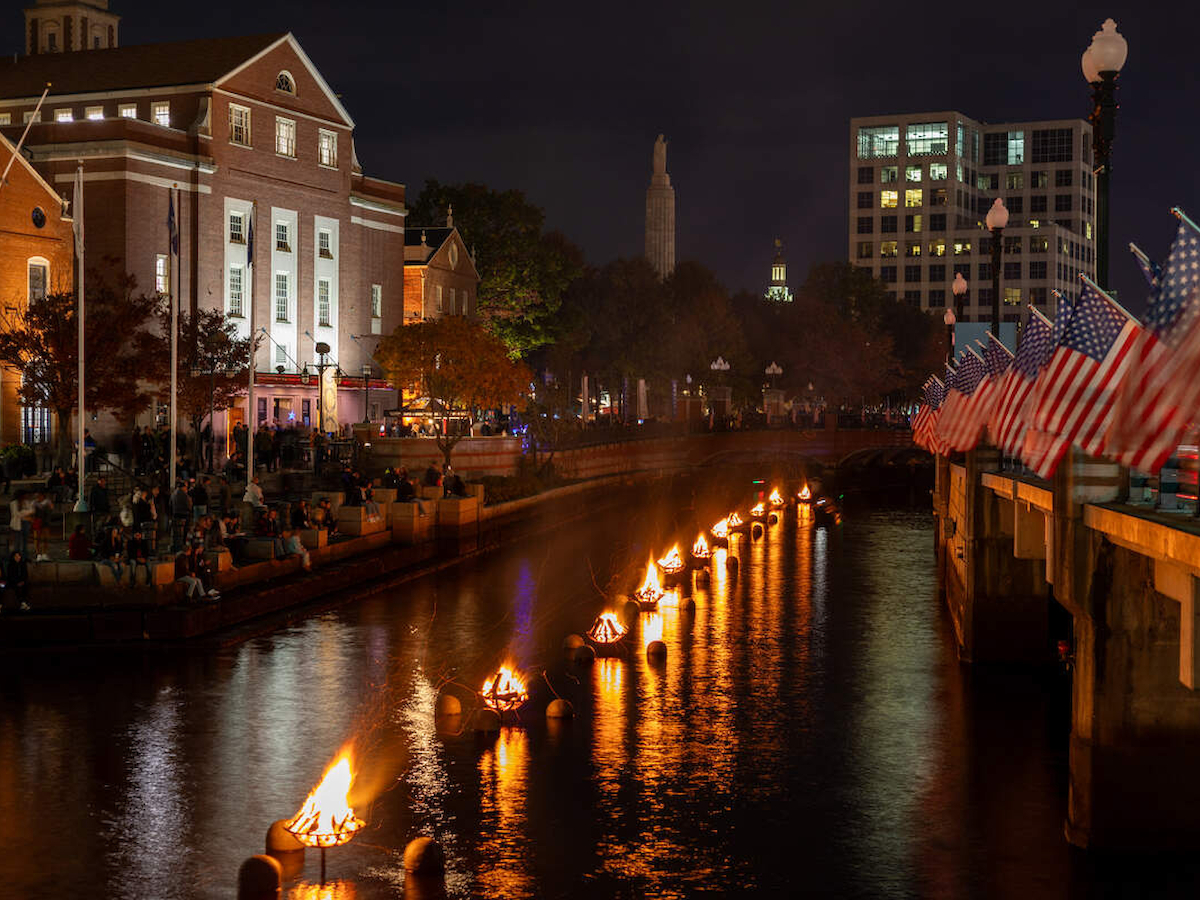 A nighttime scene features a canal with floating fires, buildings, and American flags along the water, creating a vibrant city atmosphere.