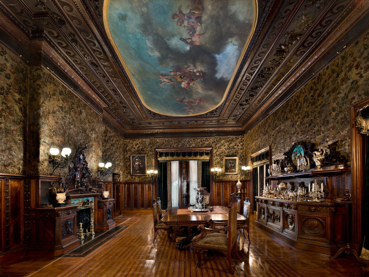 The image shows an opulent room with ornate wooden furnishings, a painted ceiling, and a dining table set in the center, adorned with various decorations.