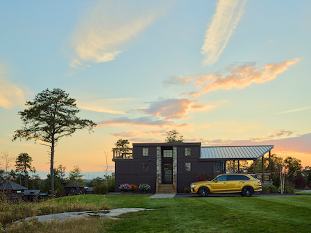 This image shows a modern house with large windows and a yellow vehicle parked outside under a partly cloudy sky, during sunset.