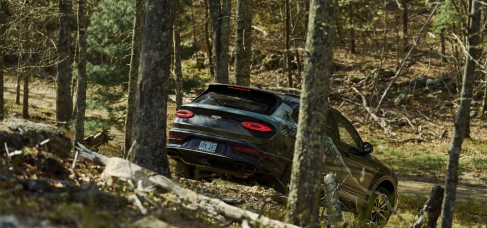 A dark-colored SUV is driving off-road through a forested area with trees and rough terrain in the background.