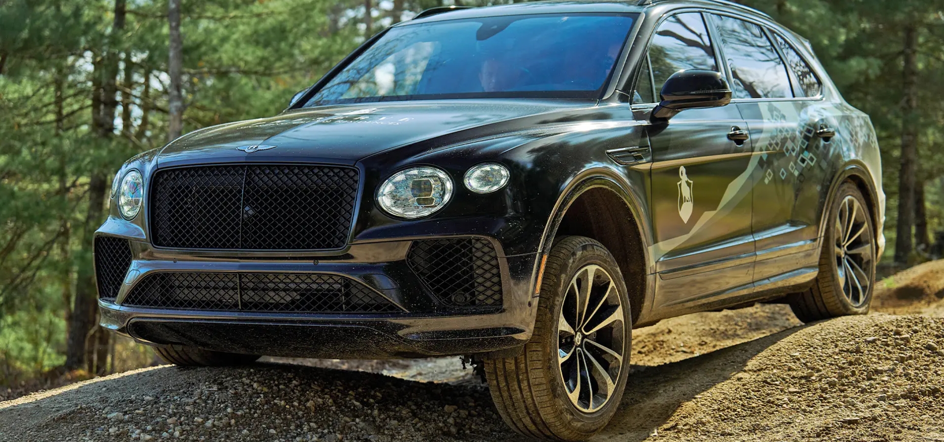 Black Bentley Bentayga driving on a dirt road surrounded by trees.