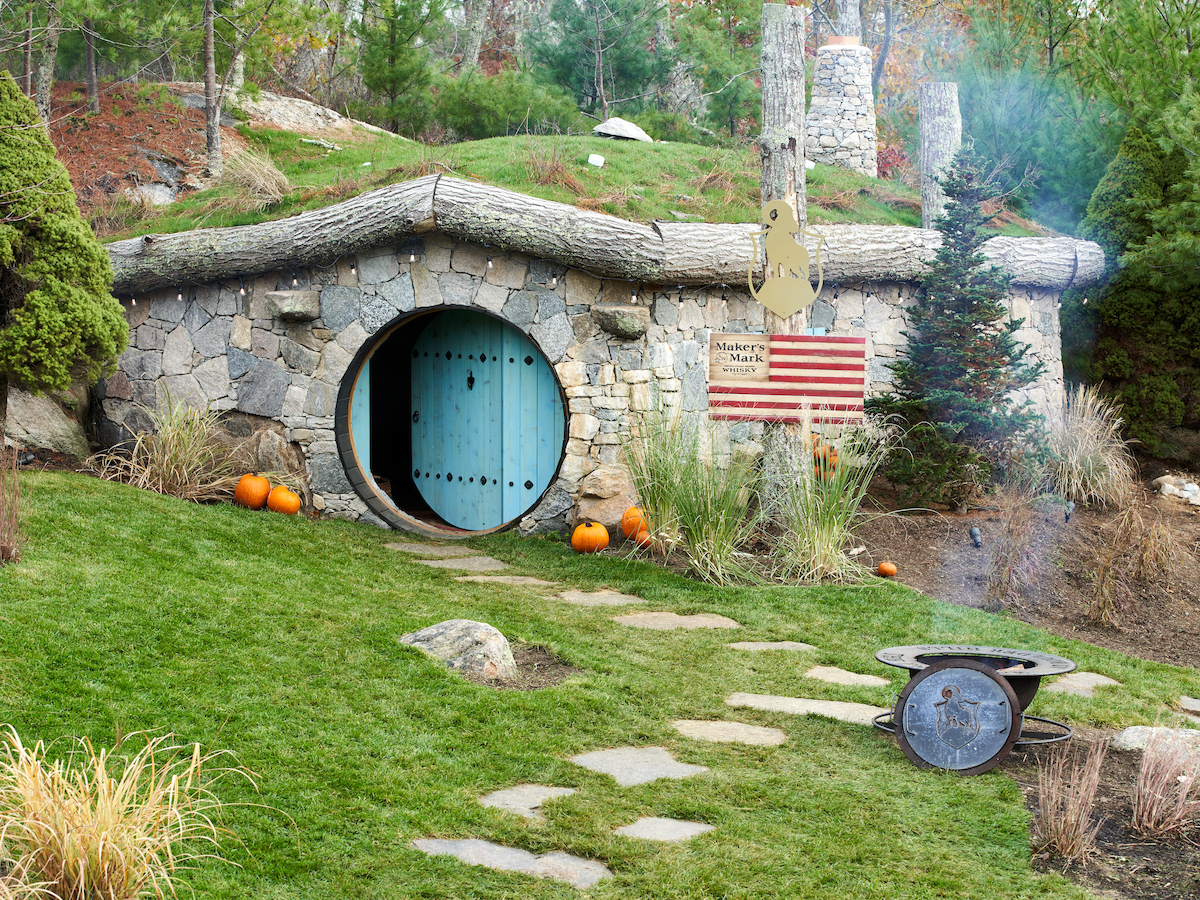 A stone-built house with a circular blue door, surrounded by greenery, pumpkins, a path, and an American flag. Smoke is seen on the right.