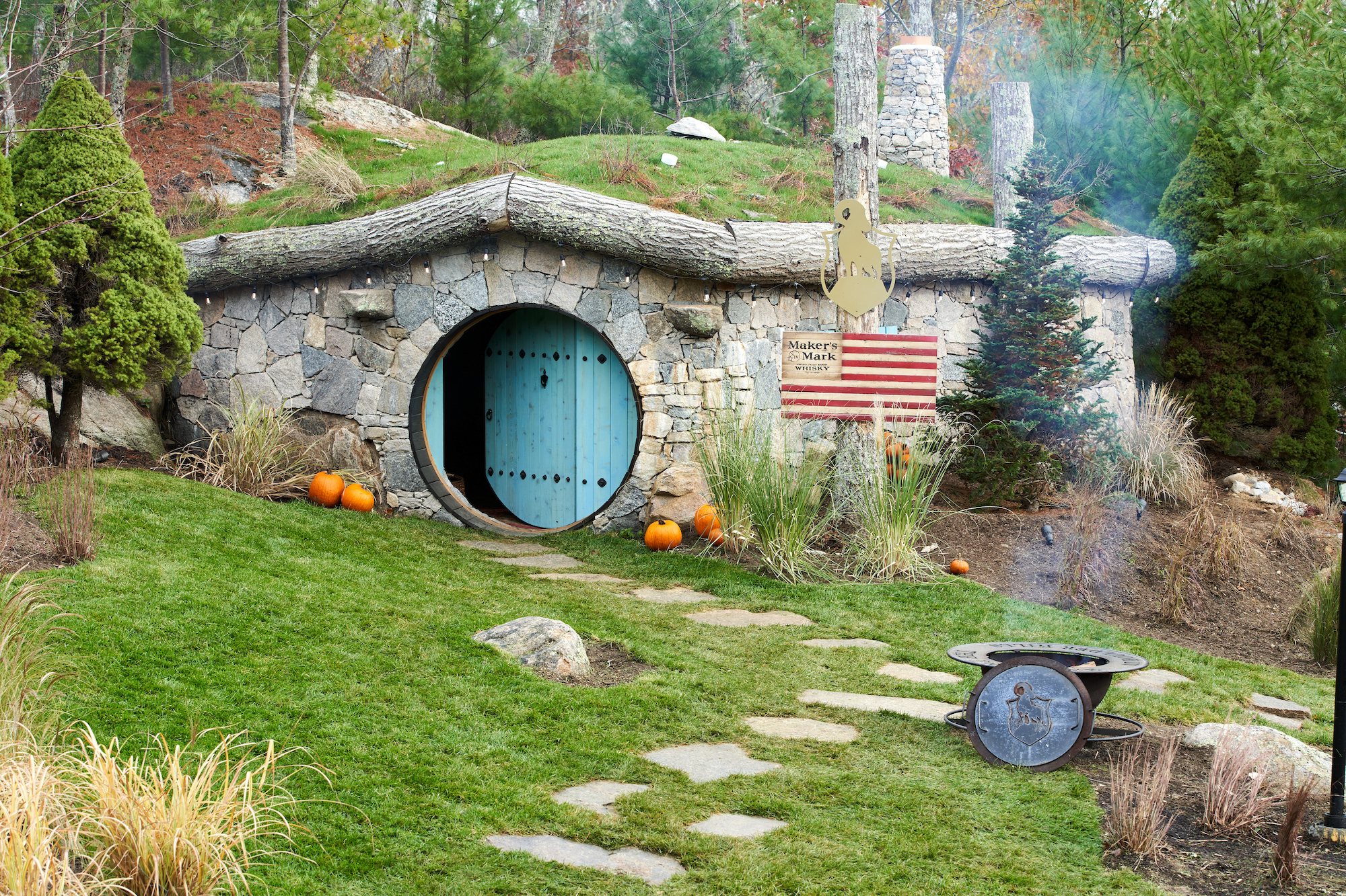 A Hobbit-like home with a circular blue door set in a hillside, surrounded by greenery, pumpkins, and an American flag on the wall.