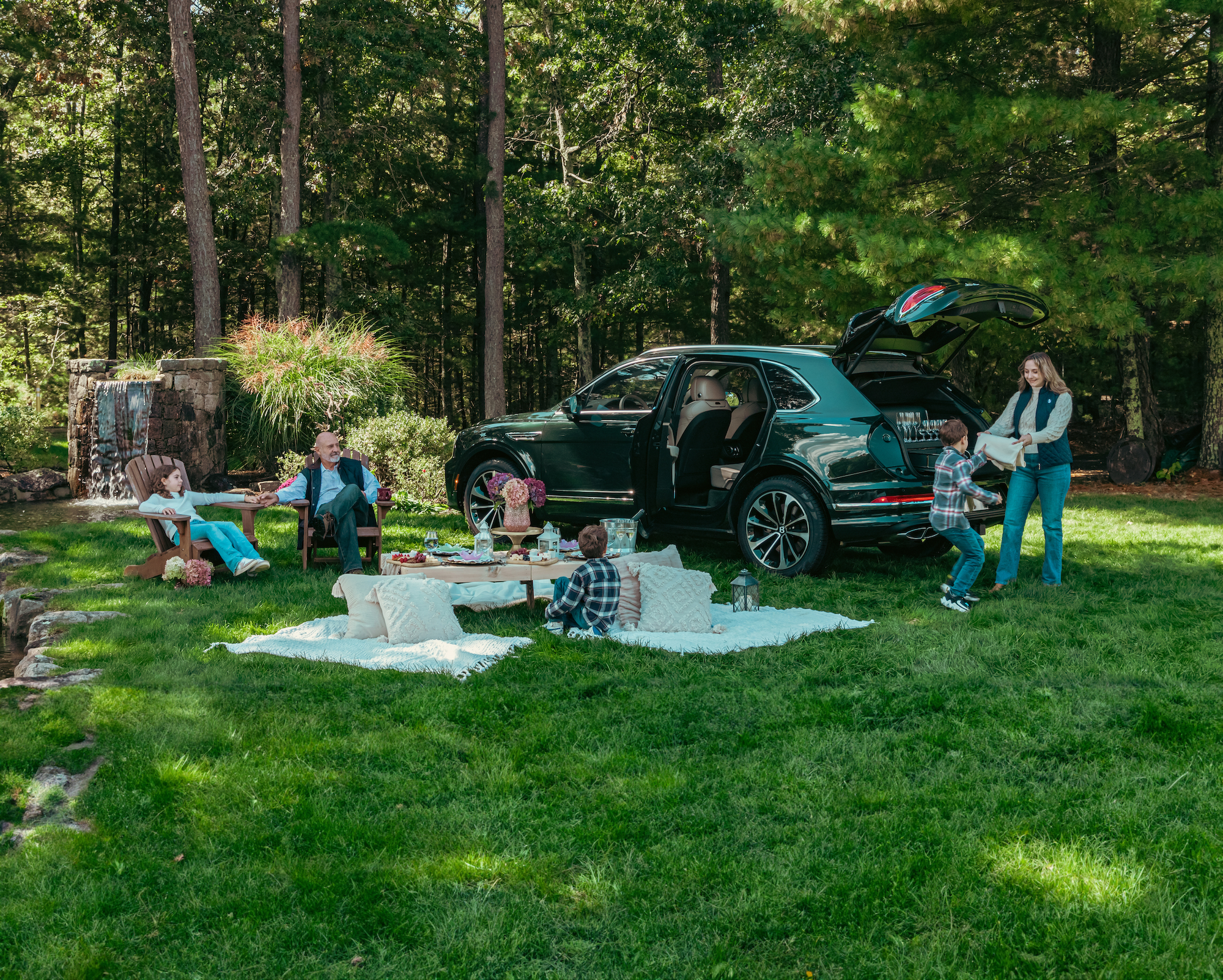 A group of people are having a picnic near a black SUV in a forested area, with kids playing and adults setting up and relaxing.