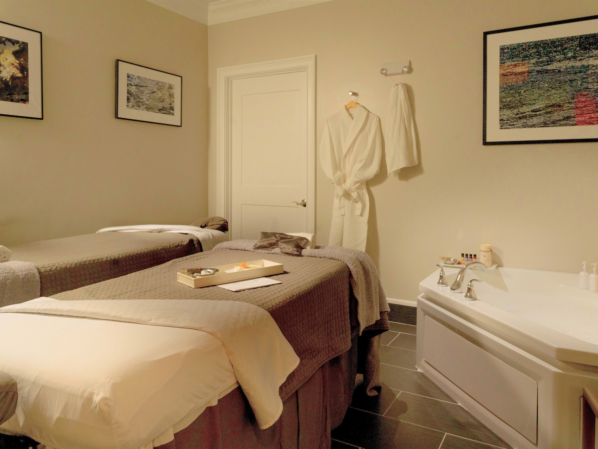 The image shows a serene spa room with two massage tables, a bathtub, a robe hanging on the wall, and framed artwork, exuding relaxation.