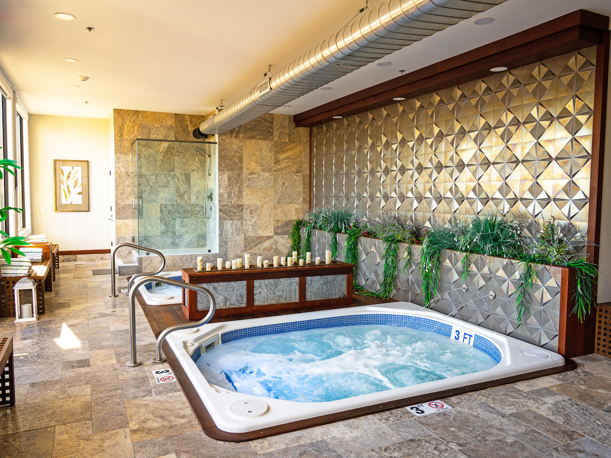 This image shows an indoor spa area with a hot tub, decorative wall, plants, benches, and candles, creating a relaxing ambiance.