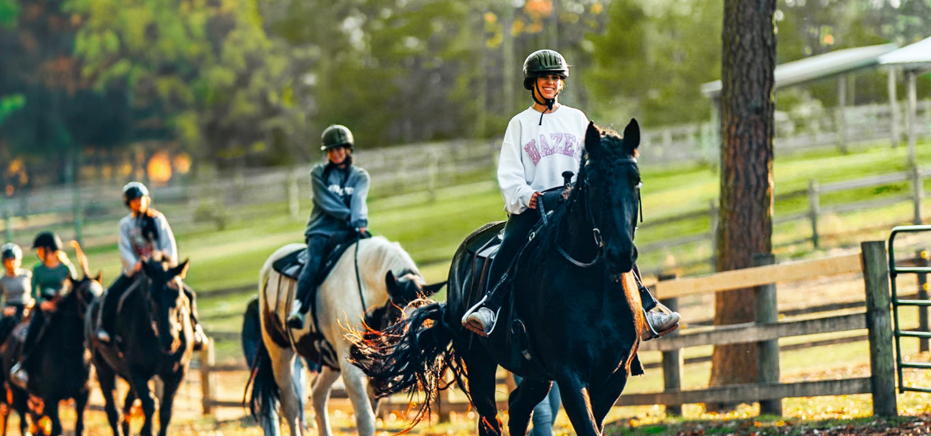 People are riding horses along a trail, wearing helmets and casual clothes, with a wooden fence and trees in the background.