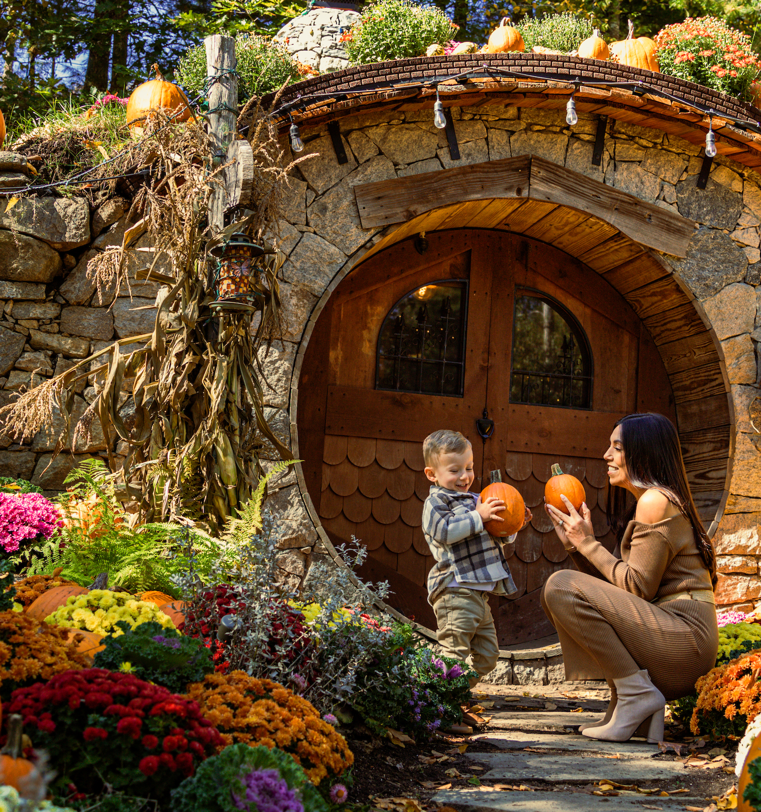 A woman and child are outside a hobbit-like house decorated with pumpkins and flowers, enjoying a festive autumn day.