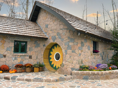 A stone cottage with a rounded door featuring a sunflower design, surrounded by potted plants and pumpkins, and a flagstone pathway.