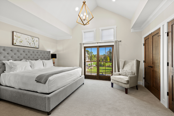 This image shows a modern bedroom with a large bed, an upholstered headboard, a chair, and glass doors leading to an outdoor view.