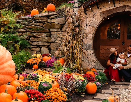 A family sits by a round doorway, surrounded by pumpkins and colorful flowers, creating a cozy autumn atmosphere.