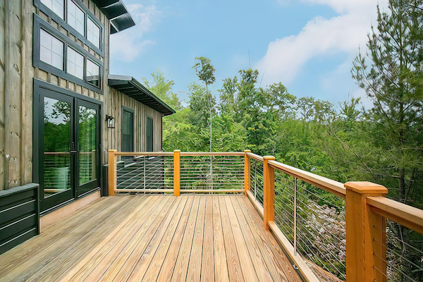 A wooden deck attached to a house with large glass doors overlooks a lush, green forest under a clear blue sky.