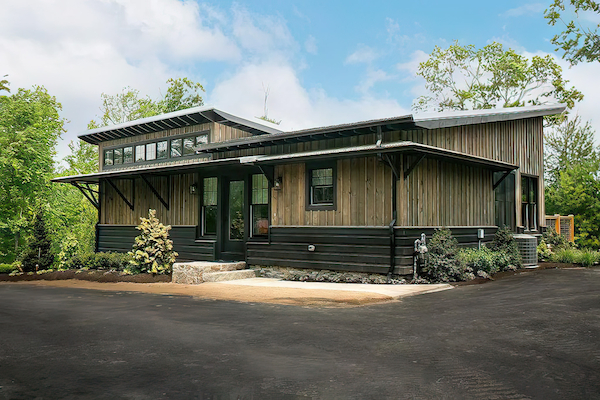 A modern, single-story house with a wooden exterior and large windows, surrounded by trees and a paved driveway.