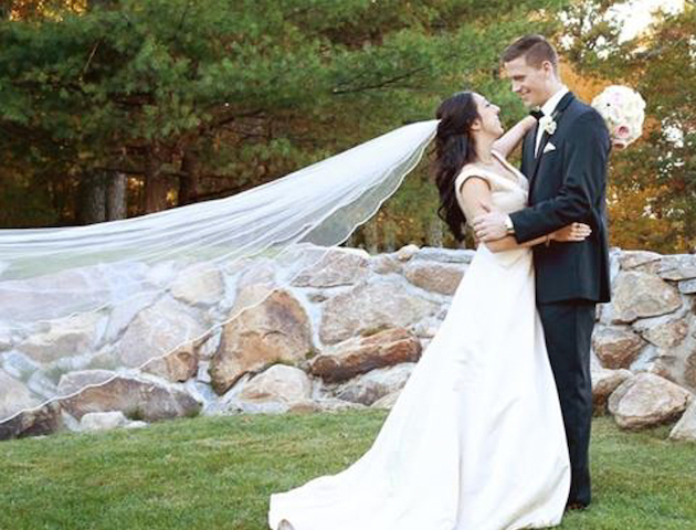 A bride and groom stand on grass, embracing in front of rocks and trees, with the bride's veil flowing in the breeze.