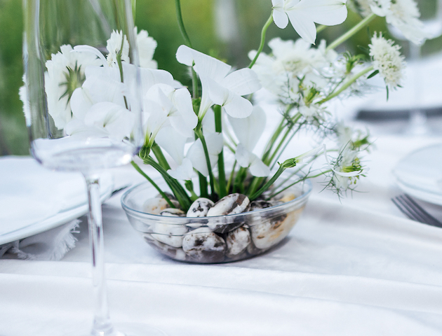 A table set with elegant white flowers in a glass vase, white plates, and wine glasses on a smooth, white tablecloth.