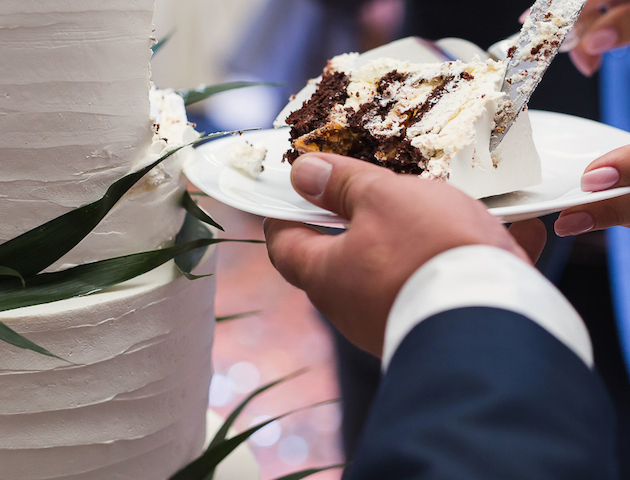 A person is serving a slice of cake from a tiered, white frosted cake decorated with green leaves onto a plate.