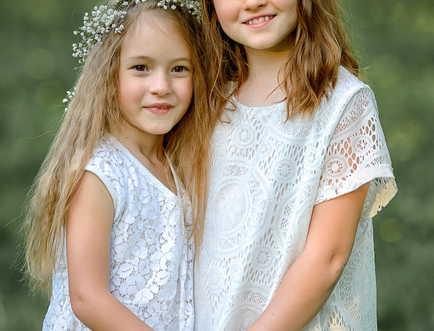 Two children in white dresses stand in a grassy area, wearing floral wreaths on their heads, holding hands, and smiling at the camera.